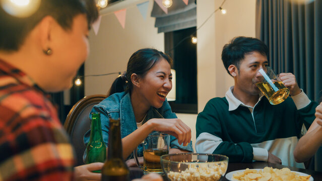 Group of multiethnic adult Asia friend drink alcohol beer bottle glass and fun talk laugh enjoy hangout party at dining table in night life indoor home. Happy hour millennial generation inside house.