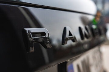 Obraz premium Close view of Range Rover rear letters in focus, glossy black paint reflects light. Soft blur hides other letters in a well lit workshop with luxury models nearby.