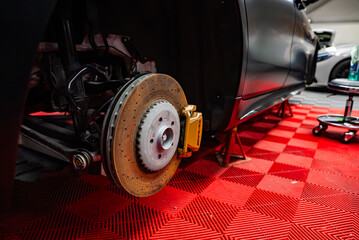 Close up of a high performance car on jack stands in a workshop. A drilled rotor and gold multi piston caliper are exposed, with red checkered floor and tools visible.