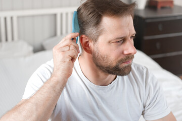 Handsome man combing brushing hair sitting at home light white bedroom. Guy styling grooming his...