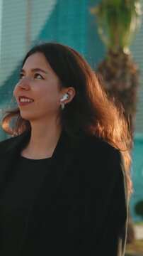 Cheerful young businesswoman walking through a bustling city street, smiling with wireless earbuds, soaking up sunlight between skyscrapers while commuting or taking a joyful break