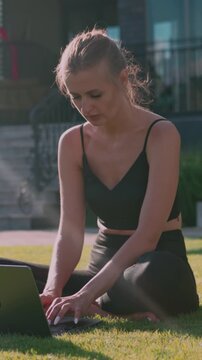 Young athletic woman is seated on a lush green lawn, typing on her laptop with focused determination in a peaceful outdoor environment bathed in gentle sunlight