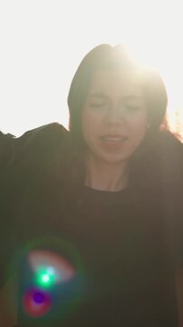 Lively young woman with flowing dark hair dancing and singing on a sunlit city street at sunset, carefree and joyful, lost in music with wireless earbuds and radiant lens flare