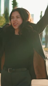 Brunette woman in a stylish black business suit twirling on a sunny city street at sunset, smiling freely and celebrating carefree urban joy and lively evening motion