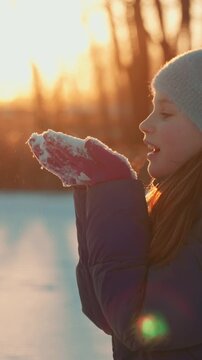 Joyful little girl in cozy winter clothes playing in snow at a sunlit park, blowing flakes and enjoying a warm, festive sunset that adds magical holiday charm to the scene
