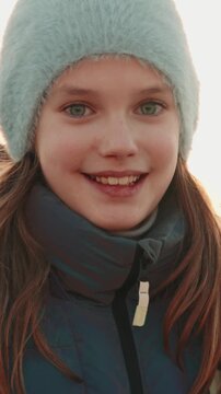 Charming close-up of a smiling young girl with green eyes in a cozy hat and jacket, basking in warm sunlight and lens flare while enjoying an outdoor winter day