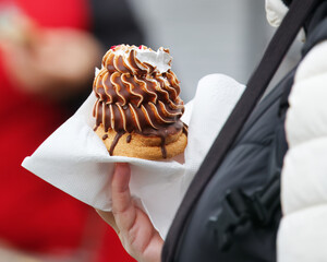 A visitor at the Naplavka market holds dessert locally called inidan, light sponge base topped with a tall swirl of chocolate-coated whipped foam, glossy drips and a delicate whipped-cream accent.