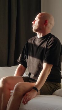 Vertical video of a man waking up in a softly lit bedroom, removing the blanket and stretching while sitting on the bed. A calm morning routine moment at home. High quality 4k footage
