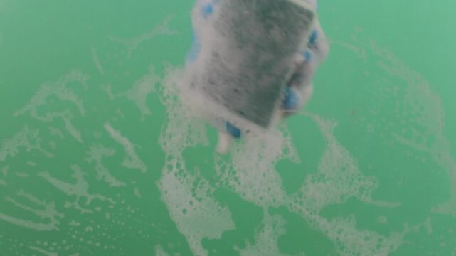 A hand in a rubber glove with a sponge washes the surface. Hands in rubber gloves hold the sponge and scrub the surface. The sponge is covered in sudsy cleanser on a chroma key.