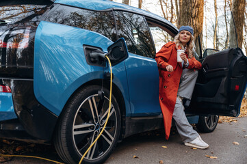 Automobile is charging. Beautiful blonde woman is with her blue electric car on the road of forest