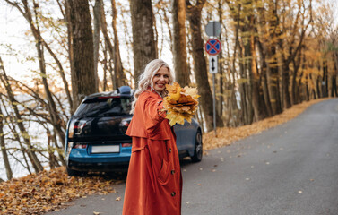 Bunch of fallen leaves, fun. Beautiful blonde woman is with her blue electric car on the road of forest