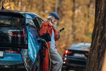 Black smartphone in hands, standing and waiting for charging to complete. Beautiful blonde woman is with her blue electric car on the road of forest