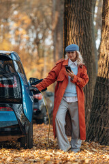 Rural non urban scene. Waiting for auto to charge. Beautiful blonde woman is with her blue electric car on the road of forest