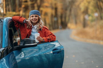 Smiling, having fun. Beautiful blonde woman is with her blue electric car on the road of forest