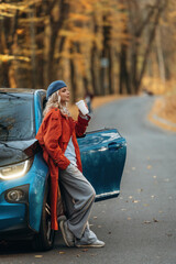 Enjoying the drink, leaning on automobile. Beautiful blonde woman is with her blue electric car on the road of forest