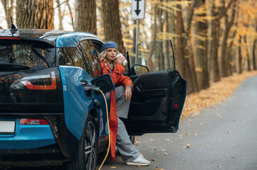 Process of charging. Beautiful blonde woman is with her blue electric car on the road of forest
