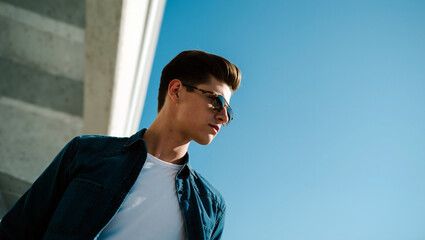 A young handsome man in a denim jacket and sunglasses stands confidently against a concrete wall, enjoying the sunny weather with a clear blue sky. Generative AI.