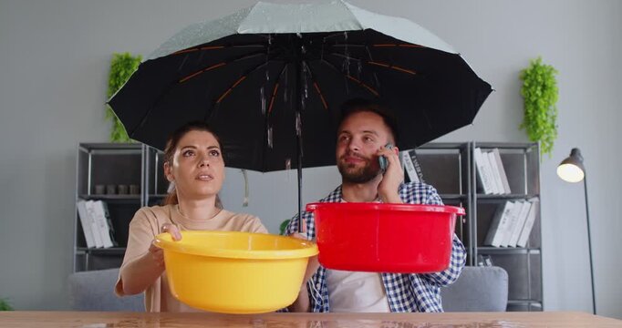 Couple with umbrella holding buckets under ceiling leak. At home, they catch water while calling for repair to reduce damage and stains. Leakage, home maintenance and emergency response.