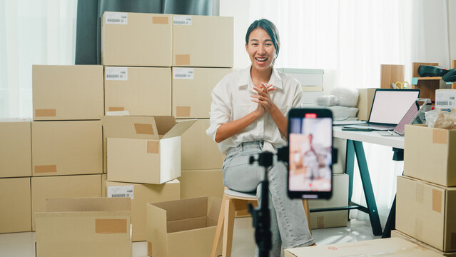 Youth Asia lady owner small business online shop sit front of phone look at camera live streaming talk to customer order promotion with full carton box at home. Shipping and delivery concept.