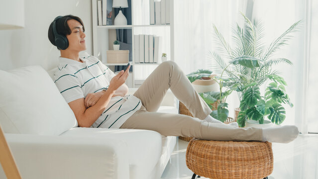 Youth attractive Asia male with casual cloth calm feeling lay down on sofa wear headphone hold smartphone listen music playlist relax at living room in cozy house. Lifestyle leisure at home concept.