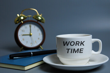 Desk setup with coffee cup, notebook, and pen, blurred clock in background, “WORK TIME” displayed, symbolizing starting daily tasks.