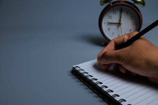 A calm scene showing a hand writing in a spiral notebook with a classic alarm clock behind, symbolizing self-care time, mindful journaling, and taking moments to pause for personal wellness.