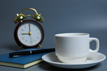 White coffee cup on a saucer beside a notebook and pen, with a blurred alarm clock in the background, symbolizing focus, planning, productivity, time management, and a calm work routine.