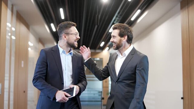 Two smiling businessmen engaging in casual discussion while walking along corridor of businss center. Happy colleagues have friendly conversation, talking. Teamwork, communication, collaboration