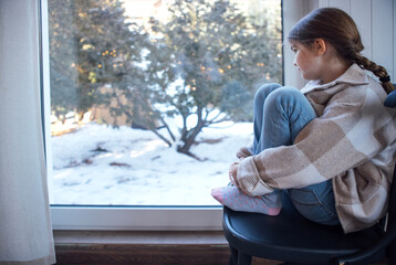 Young girl sitting on a chair, gazing out a window at a snowy landscape, wearing a cozy sweater, reflecting on winter's beauty and tranquility in a serene indoor environment