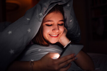 Young girl with brlond hair, smiling while using smartphone under cozy blanket, illuminated by screen light, creating a warm and inviting nighttime atmosphere for relaxation