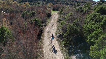 A drone follows an athletic man wearing a tank top, shorts, and a trail running backpack as he runs along a mountain trail on a sunny day. Conceptually, it's a healthy and active lifestyle.