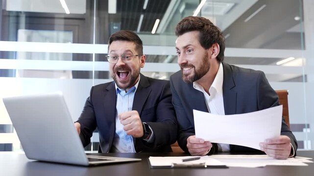 Two happy businessmen celebrate successful results using laptop while sitting at desk in modern business office. Joyful excited colleagues show joy and excitement while reading great news on computer