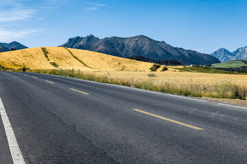 Empty asphalt road running alongside a golden hillside with a majestic mountain range in the background on a sunny day.