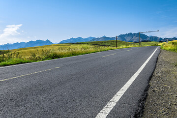 Empty asphalt road on a hillside with green grass and a mountain range on the horizon under a blue sky.