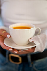 A person is holding a white cup filled with an orange beverage on a matching saucer. The focus is on the drink and hands with white manicured nails