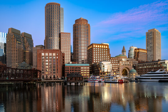 A stunning view of the Boston downtown skyline at twilight, seen across the water from the Seaport District, with brightly lit towers and colorful reflections.
