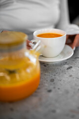 A person is holding a white cup of vibrant orange sea buckthorn tea, served with a matching teapot on a modern grey surface in natural light