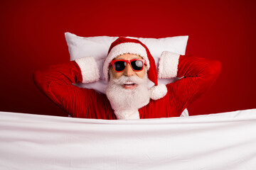 Obraz premium Cheerful Santa Claus lying in bed with sunglass smiling on a festive red background, spreading holiday cheer and joy