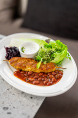 Grilled kofta kebab on a skewer served on a white plate with kidney bean stew (rajma), fresh green salad, pickled red cabbage, and a side of yogurt dip
