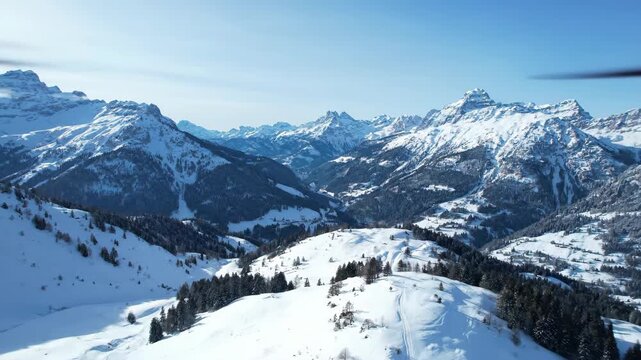 Aerial vista of snow-covered mountain range under clear blue sky. Rugged peaks and slopes blanketed with fresh snow and some trees