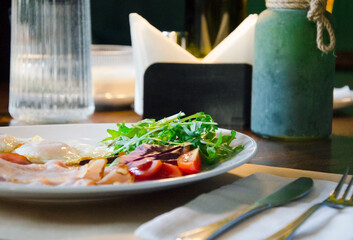 Classic breakfast plate featuring fried eggs, crispy bacon, fresh greens, and sliced tomatoes served on a white dish.