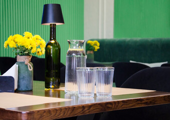 A stylish caf&eacute; table set with a glass water carafe, two drinking glasses, and a decorative bottle lamp.