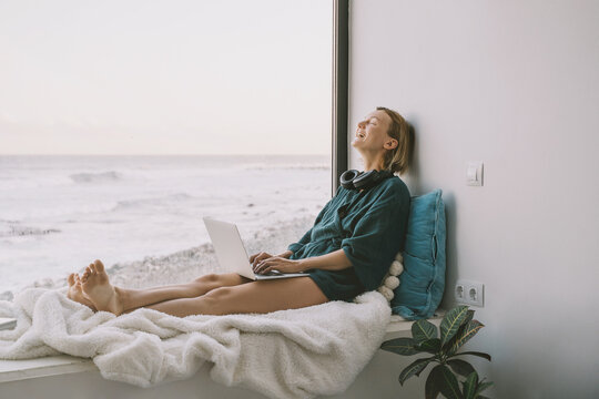 Beautiful woman using laptop by the window with sea view. Smiling woman with laptop enjoys during the weekend. Carefree female relaxes on the windowsill by large window with ocean view in the morning. - Powered by Adobe