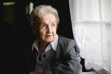 An elderly woman sits by the window, softly lit by daylight, looking outside with a calm, reflective expression as lace curtains glow beside her.