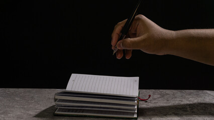 Hands holding a small notebook while writing with a black pen, captured in low-key lighting. The...