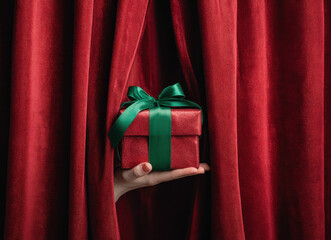 Hand holding a red gift box with green ribbon behind curtain for secret santa or holiday surprise theme