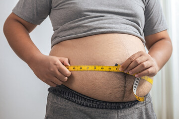 Fat or obese man with belly fat is using a tape measure to measure his waist circumference at home. He is on diet and losing weight