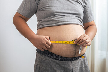 Fat or obese man with belly fat is using a tape measure to measure his waist circumference at home. He is on diet and losing weight