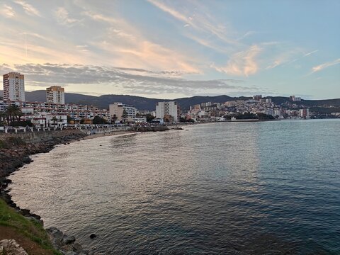 A scenic view of the coastal city of Annaba, Algeria, featuring high-rise buildings and clustered homes nestled against the hills, overlooking the calm Mediterranean Sea at sunset.