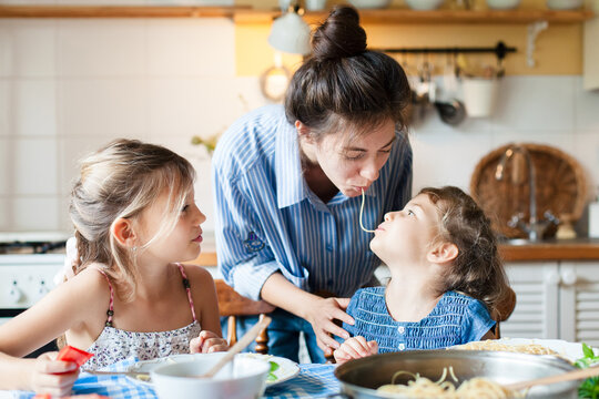 Happy mother and daughters eating pasta together at home kitchen. Playful children having fun, sharing spaghetti kiss. Kids enjoying family homemade lunch, healthy food. Candid, authentic lifestyle